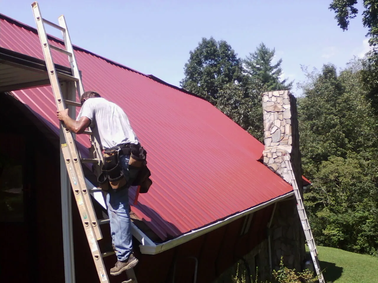 Roofer on a ladder beside a red metal roof and stone chimney — Soffit & Fascia Repair by Roofing Brothers in Green Lane